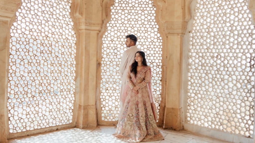 A couple in traditional ethnic wear posing against an intricate stone lattice window during a pre wedding photoshoot in a royal palace.